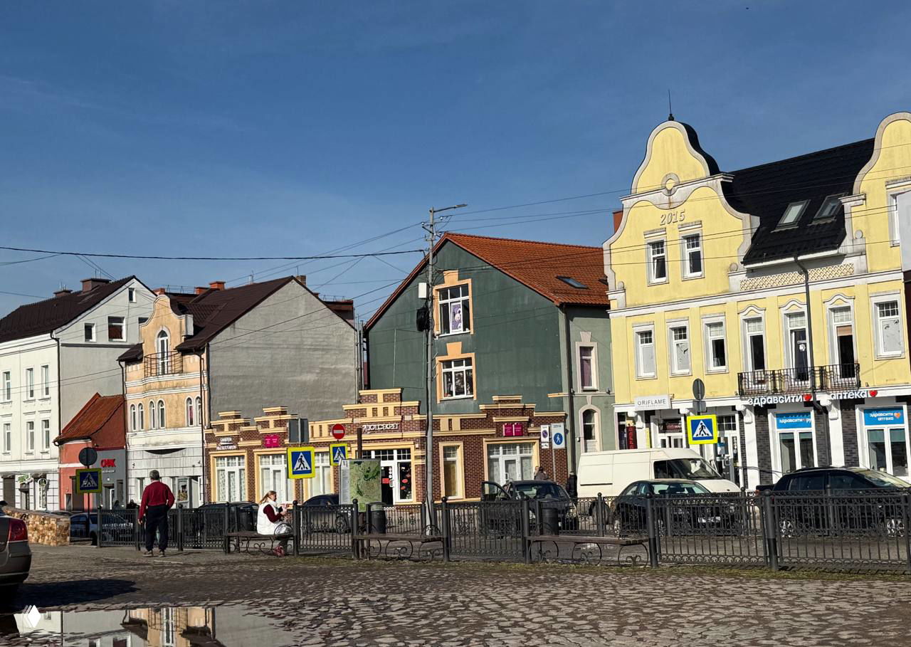 Ряд разноцветных старинных домов вдоль площади в Черняховске, люди на скамейках и машины, отражение неба в лужах на брусчатке.