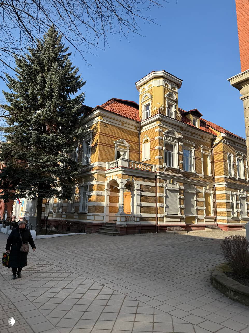 Крупное здание с башенкой и богатой лепниной в Черняховске, подъездная зона, пешеходная площадь и старые деревья на фоне синего неба.