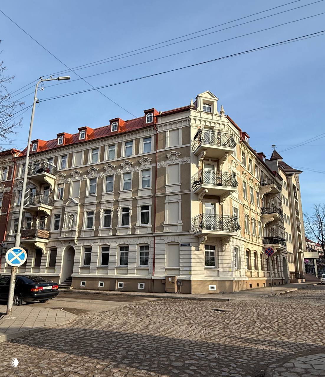 Угол многоэтажного исторического дома с балконами, декоративными фронтонами и брусчатой дорогой перед фасадом в центре Черняховска.