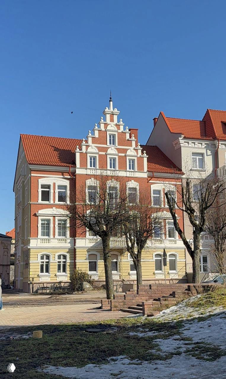 Фасад кирпичного дома с зубчатой фронтонной отделкой и голыми деревьями на переднем плане; зимне-весенний городской пейзаж Черняховска.