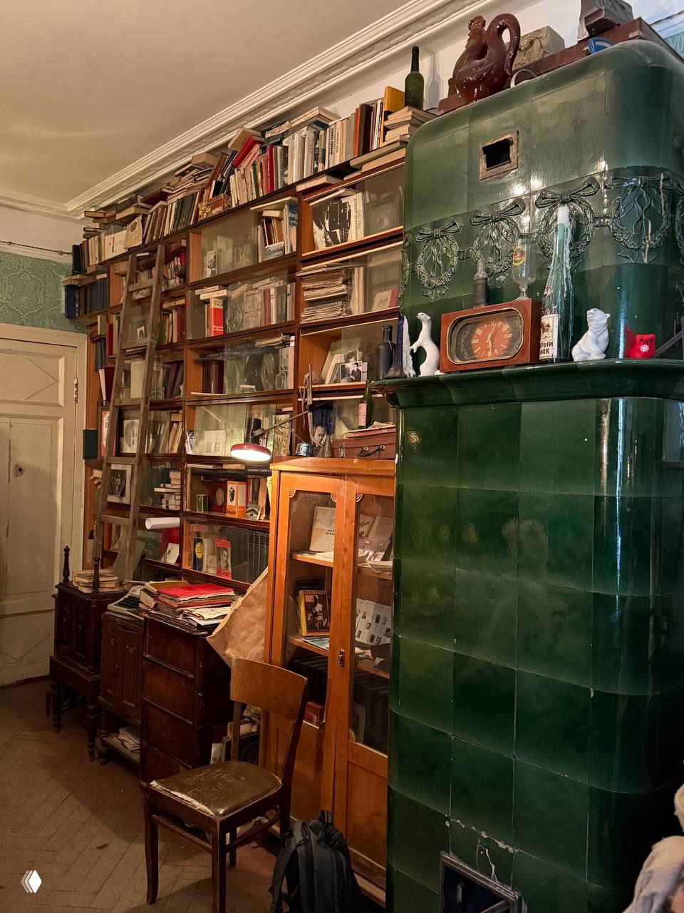 Комната с высоким книжным стеллажом, старые книги и зеленая кафельная печь рядом с креслом и рабочим столом в коммунальной квартире