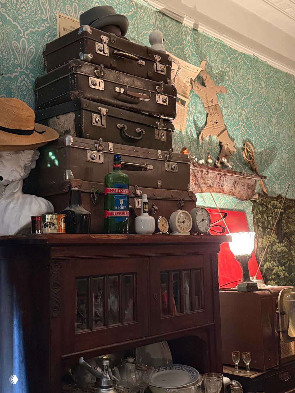 Стопка старых чемоданов и часы на шкафу, аксессуары и посуда на фоне узорных обоев в коммунальной комнате