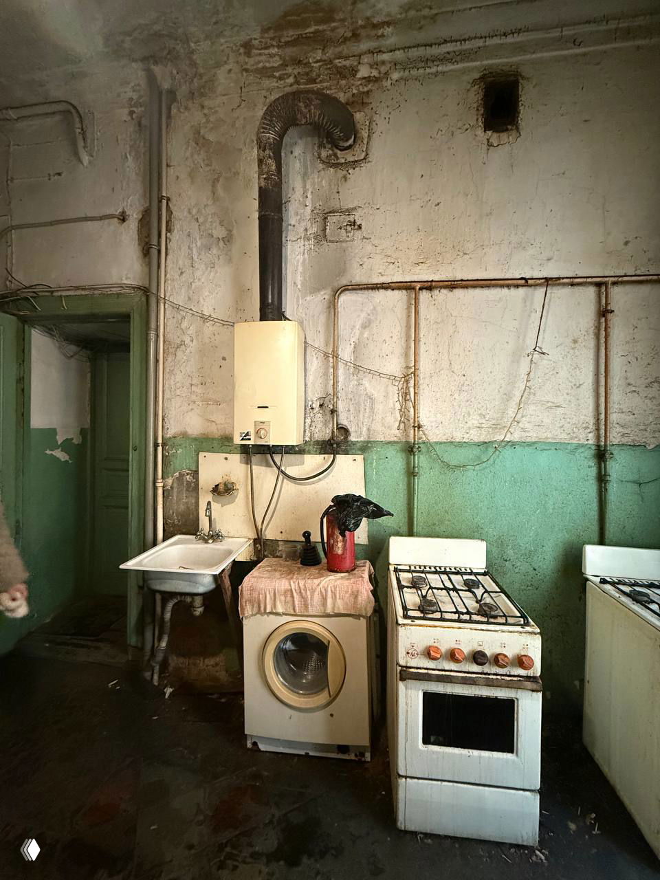 Кухонный угол с газовой плитой, стиральной машиной и мойкой в запущенной коммунальной кухне с облупившейся краской