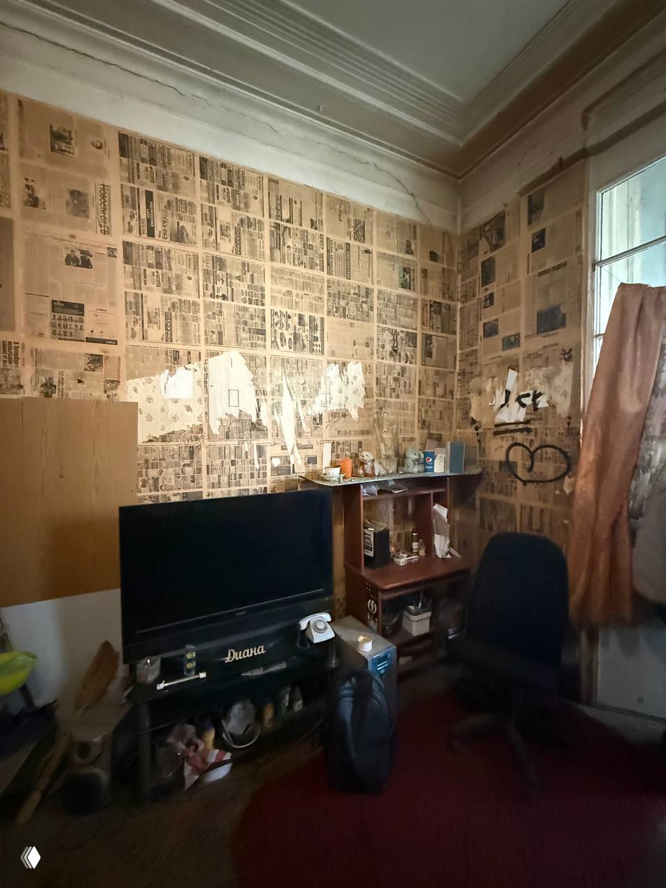 Комната с телевизором и стенами, обклеенными старой газетой, мебель и бытовая техника в плохом состоянии