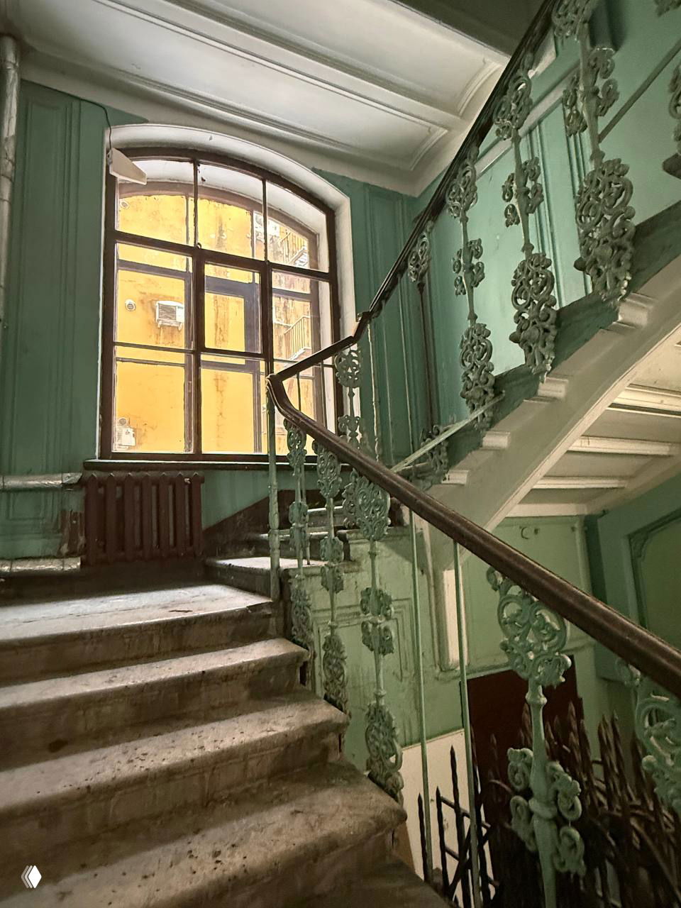 Парадная лестница старого дома с коваными перилами, естественное освещение из большого окна и потертые ступени