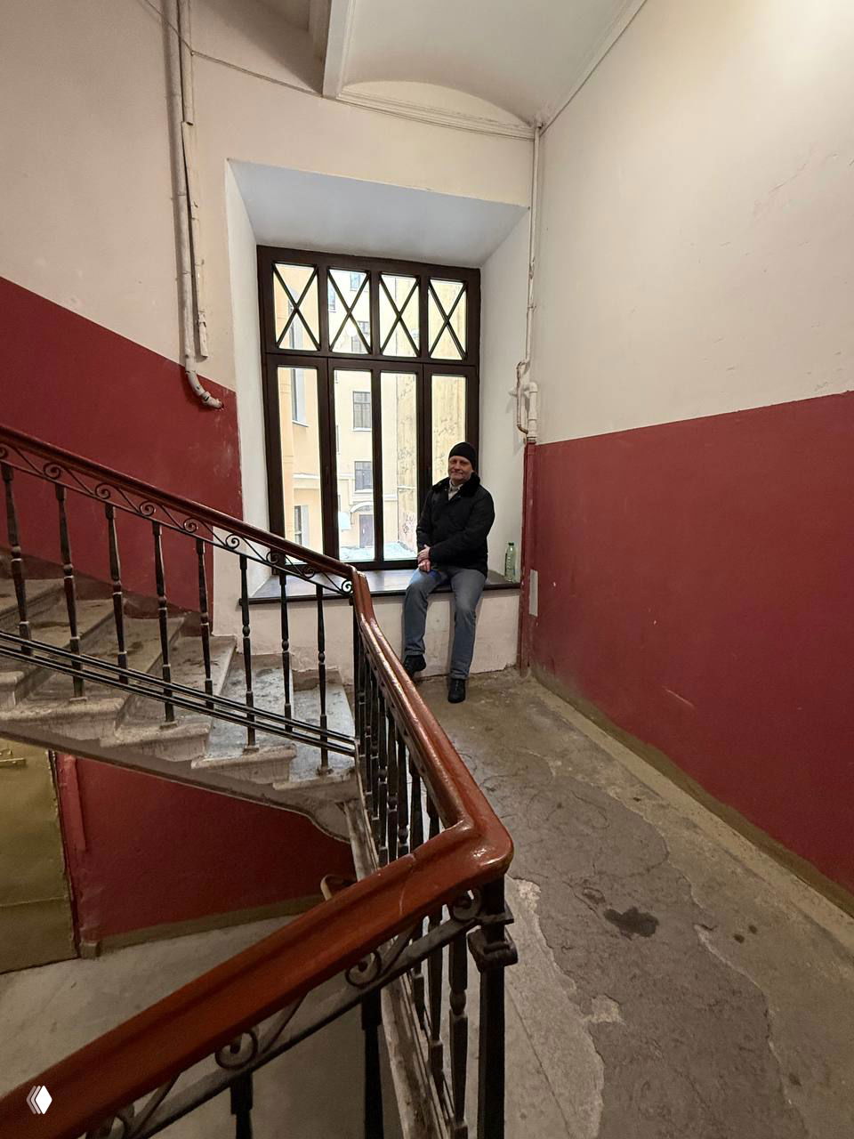 Нижняя лестничная площадка с сидящим человеком на подоконнике, простая отделка и следы износа в подъезде многоквартирного дома