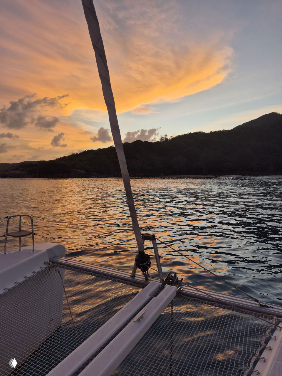 Закатный кадр с носа катамарана: тросы и сетка форштевня на фоне оранжевого неба и темного силуэта прибрежных холмов.