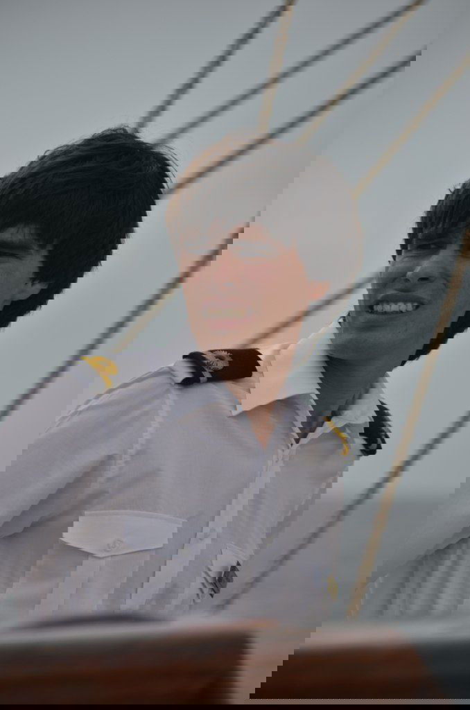 Морской офицер в белой форме с погонами на палубе парусника, видны тросы и элементы рангоута, фон — размытое море и небо.