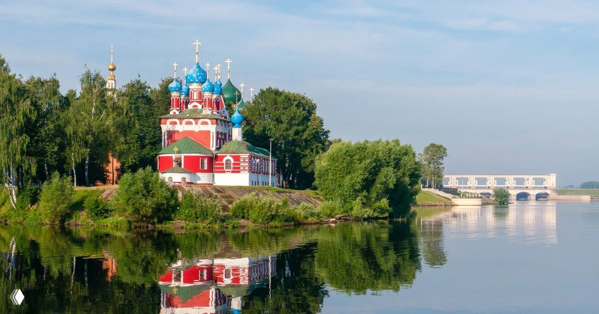 Яркая красочная деревянная или каменная церковь с синими куполами и золотыми крестами на зелёном берегу, отражение в гладкой воде.