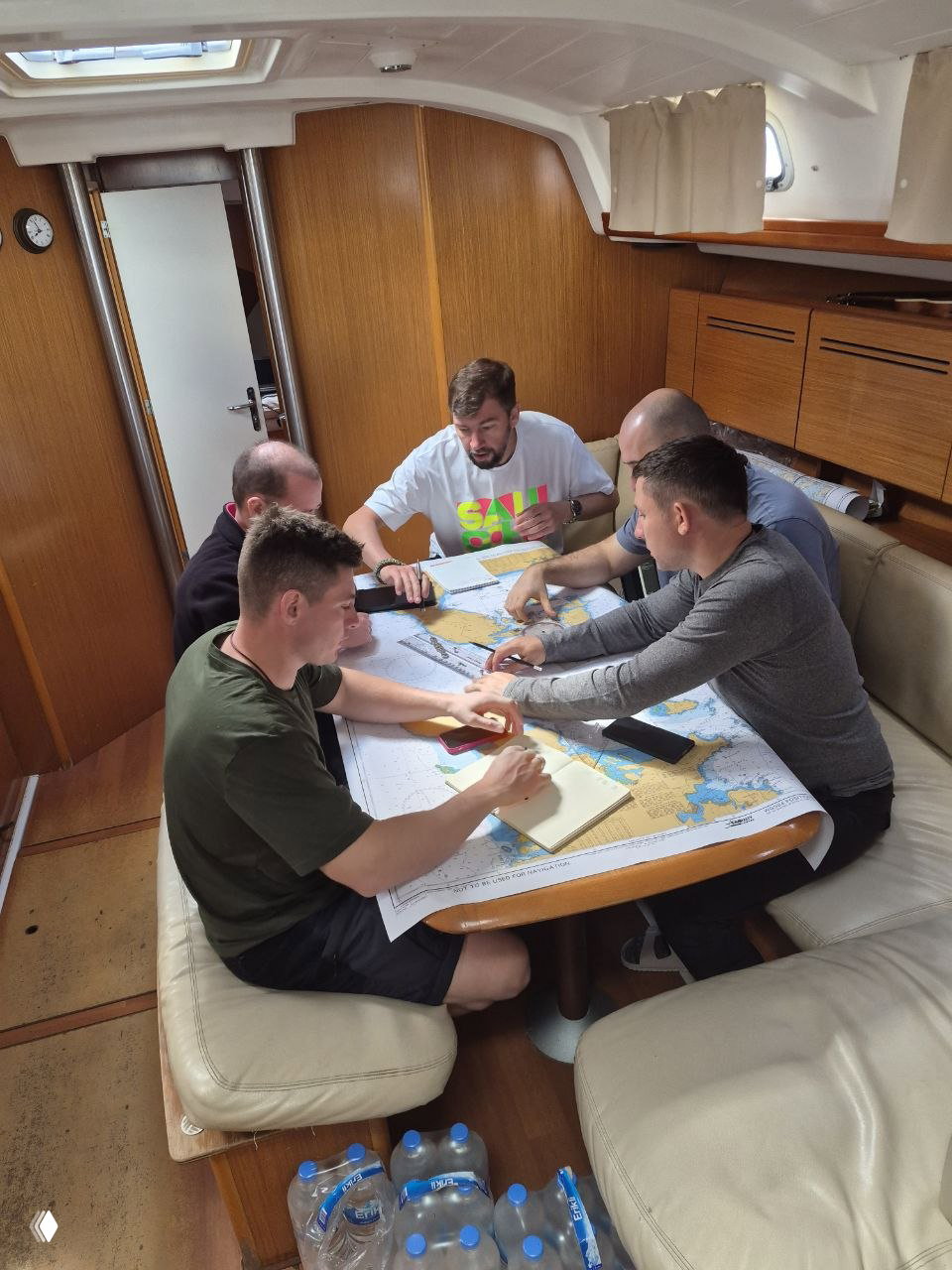 Внутри каюты группа курсантов вокруг большого морского плана на столе — коллективная работа над маршрутами, расчёты и обсуждение с инструктором.