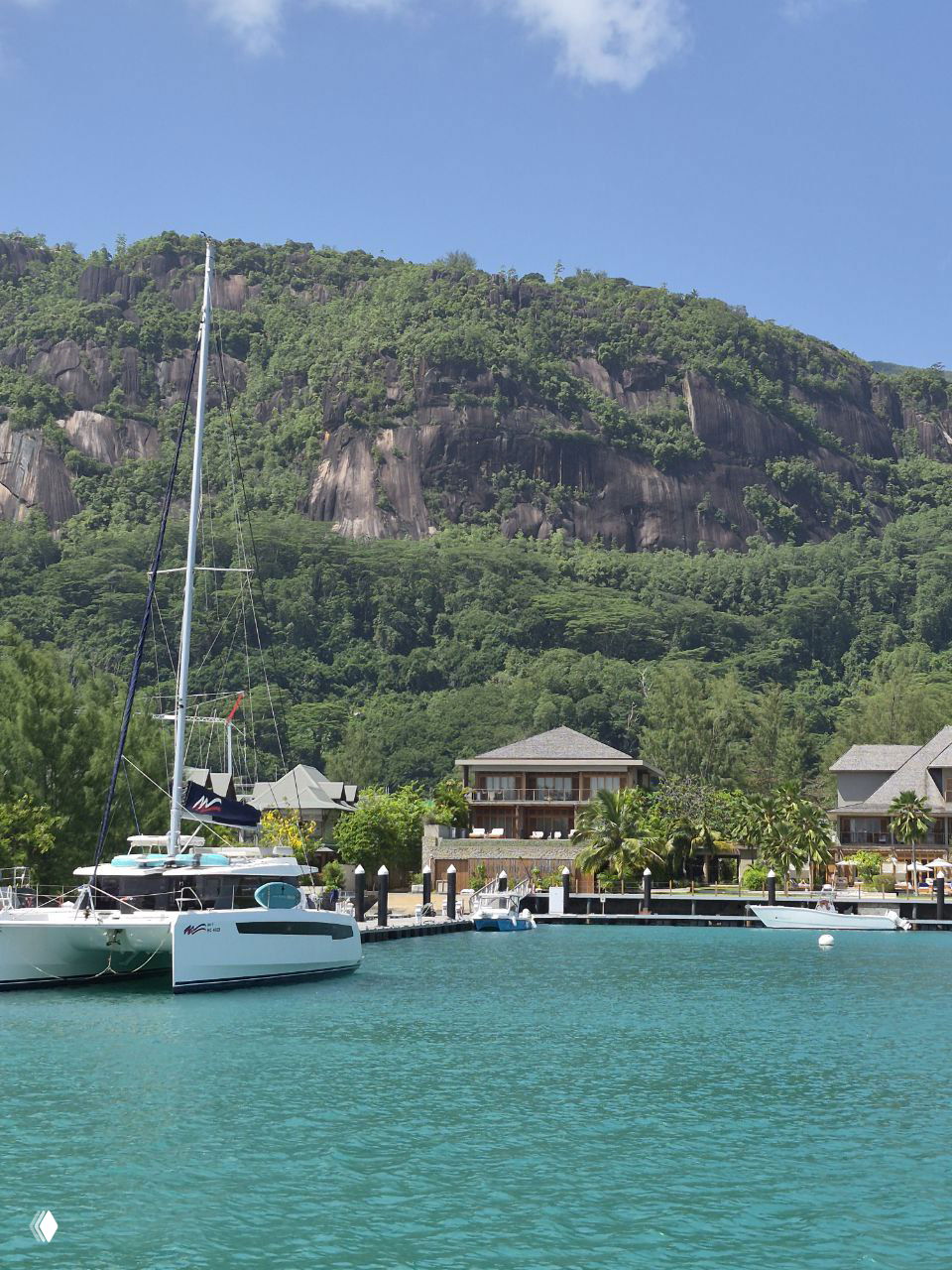 Катамаран у марина на Сейшелах на фоне гранитного холма и прибрежных вилл, бирюзовая вода и причал с яхтами.