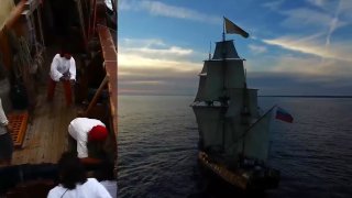Кадр из превью видео: на левой части палуба с членами экипажа, работающими на судне, справа деревянный парусный фрегат «Штандарт» под парусами на спокойном море.