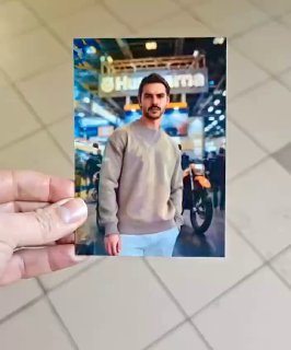 Рука держит напечатанное стерео‑фото с портретом мужчины на фоне выставочного стенда и мотоцикла; кадр сделан в зале мероприятия с плиточным полом и размытыми посетителями.