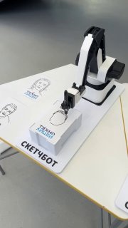 Крупный план робота-рисовальщика на столе: механизм, держатель бумаги и готовые черно-белые портреты с надписью «СКЕТЧБОТ».