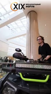 Вертикальный кадр оператора у пульта рядом с квадроциклом и роботом-рукой, люди на фоне в фойе выставки.