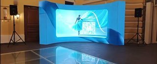 Кривой LED‑экран и светящаяся полированная поверхность на площадке мероприятия: фотозона с синим оформлением и отражением пола, демонстрация размера и формы.
