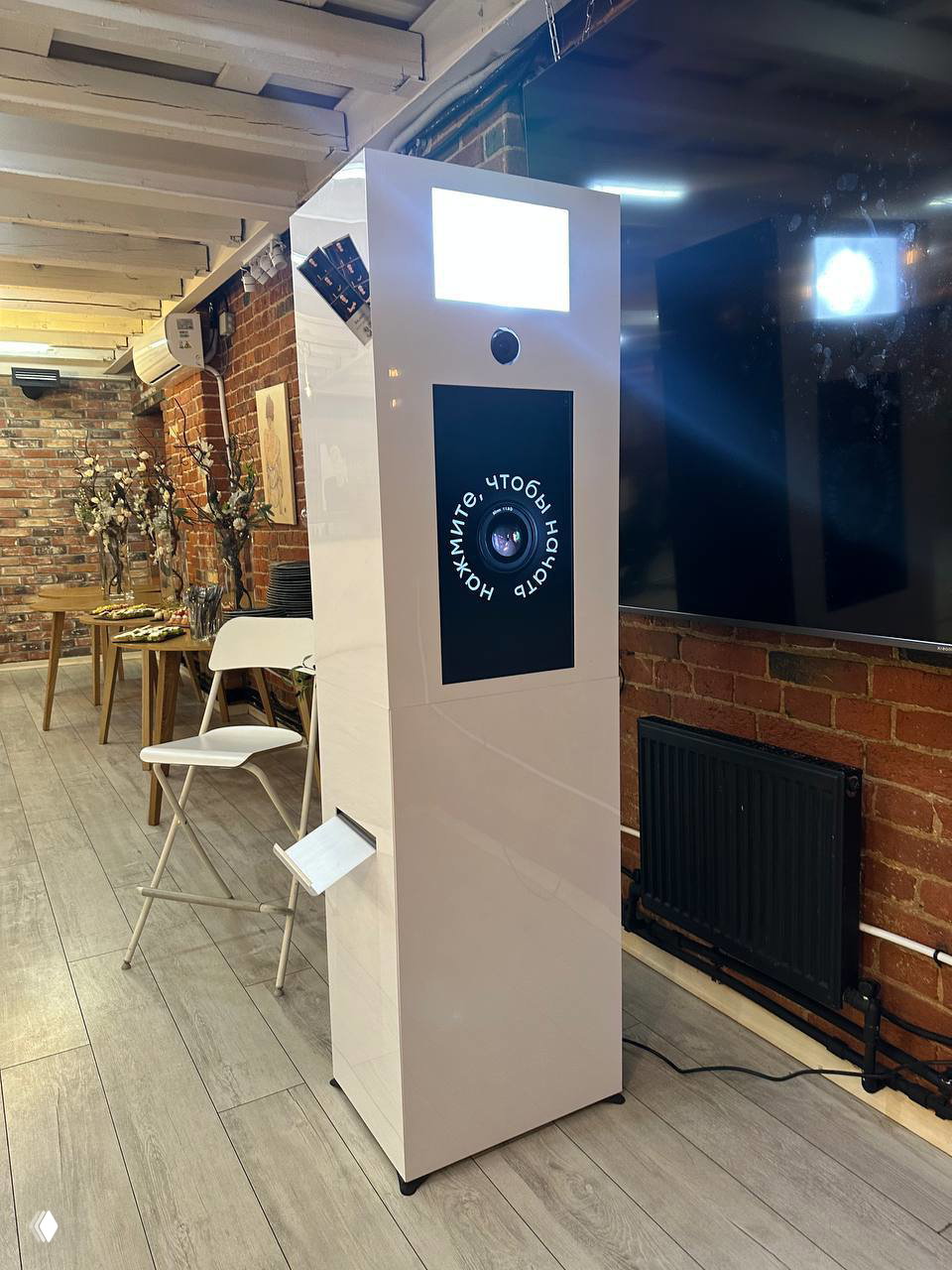 Белая глянцевая фотостойка с экраном и объективом, установлена в интерьере с кирпичной стеной и стульями — пример использования на событии или кейтеринге.