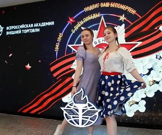 Две девушки позируют на фоне светодиодного экрана с декоративным георгиевским орнаментом и надписью, в студийной 3D‑фотозоне.