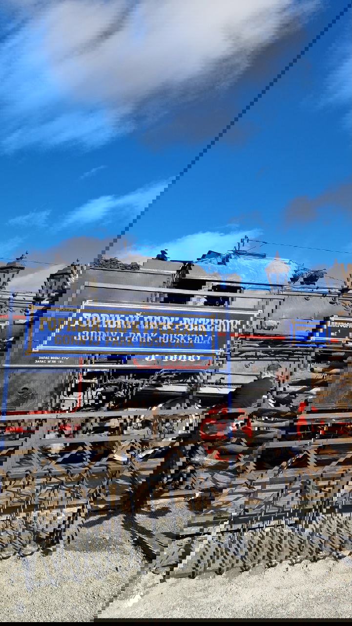 Табличка «Горный парк Рускеала» на фоне паровоза и элементов железнодорожной экспозиции: колёса локомотива, рельсы и инфраструктура парка.