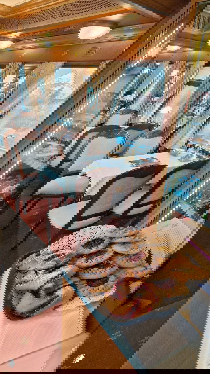 Ресторанный вагон с белыми скатертями, мягкими стульями и сервировкой; на переднем плане поднос с пирогами и выпечкой у стойки с напитками.