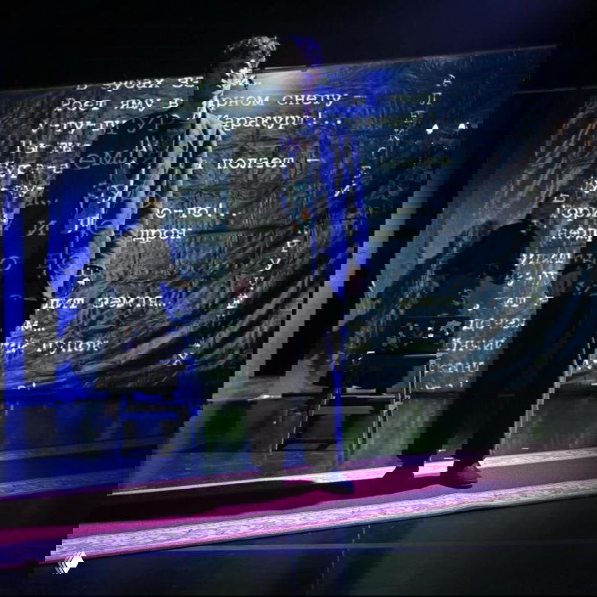 Театральная сцена в полумраке: актёр на красной дорожке на фоне проекции стихов, композиция подчёркивает поэтический характер постановки и драматическое освещение.