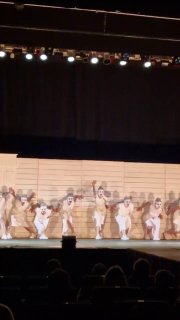 Широкий план сцены с хором актрис в белых одеждах; общее движение и колоннады декорации подчёркивают масштаб постановки.