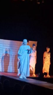 Актёр в белом костюме идёт по сцене, позади — другие исполнители; кадр подчёркивает одиночество персонажа среди хора.