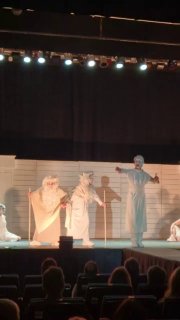 Продольный вид сцены с группой актёров в белых и бежевых костюмах; зрители видны как тёмные силуэты на переднем плане.