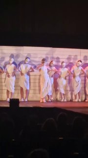 Хор актрис в белых туниках выстроен в линию на сцене; синее и тёплое освещение создают контраст и ритуальное ощущение.