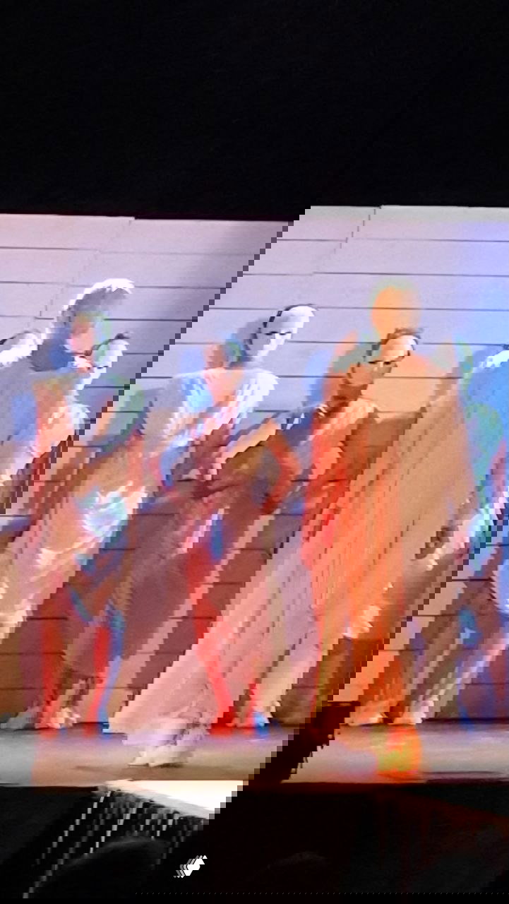 Фигуры хора в белых туниках и масках на фоне светлой декорации; строгие позы и контраст света создают ритуальную атмосферу.