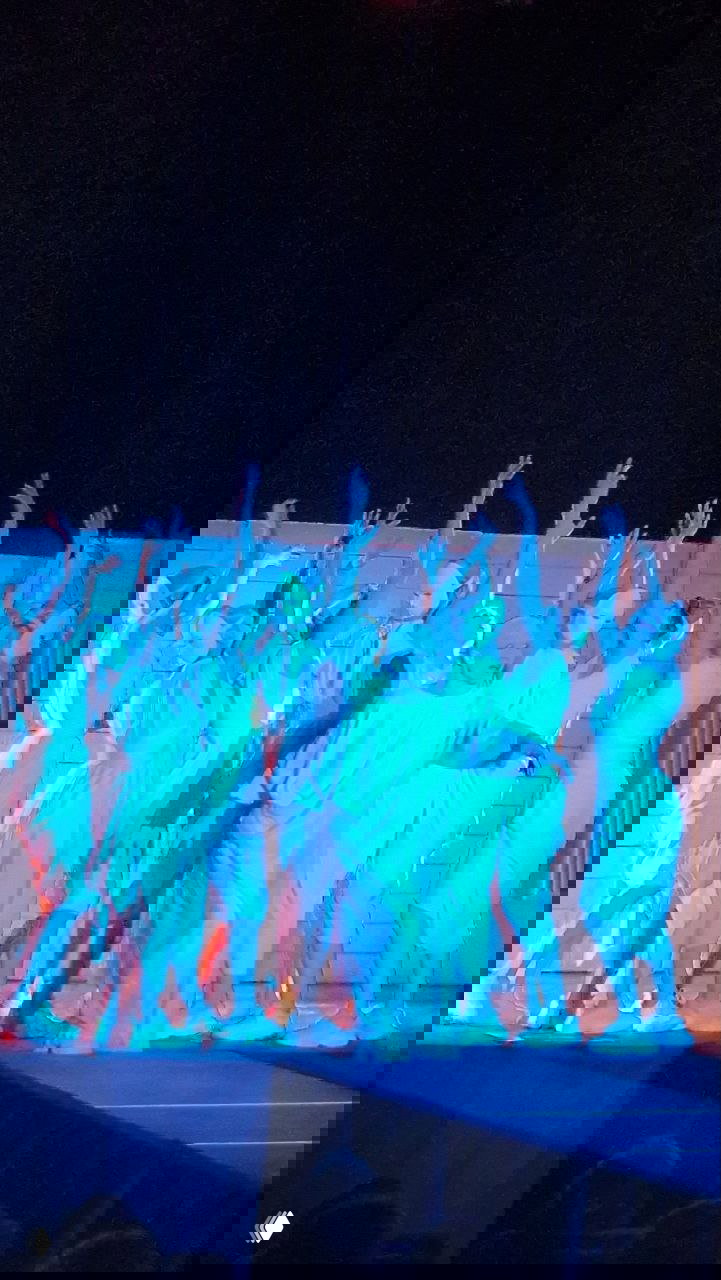 Актриса в окружении хора, освещение синий-голубое; жесты и пластика подчеркивают трансформирующую силу ритуала в спектакле.