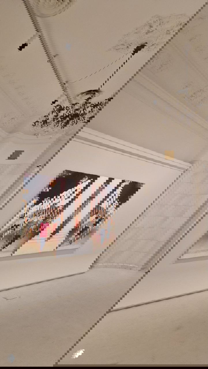 Широкий зал галереи с высокой лепниной и большой картиной на стене; пространство белых стен и парящие люстры создают музейную атмосферу