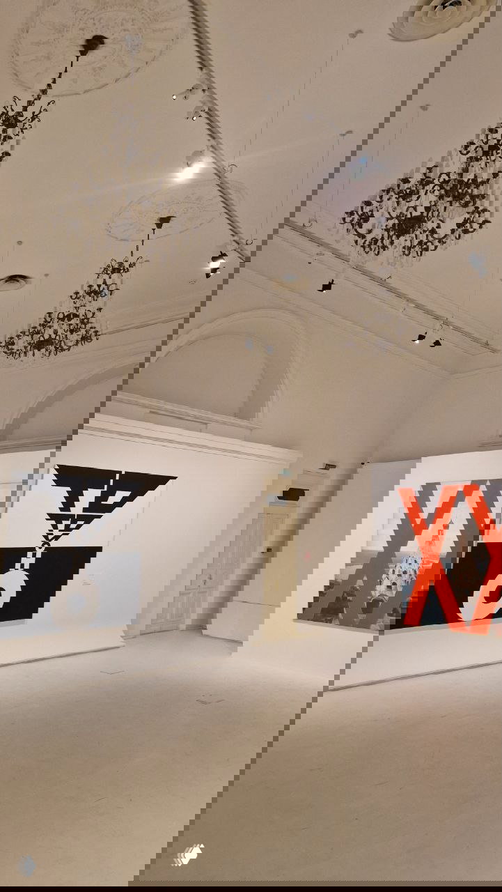 Зал выставки с двумя крупными полотнами по обе стороны стены, высокие потолки с люстрами и чистое музейное пространство вокруг работ