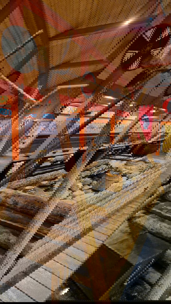 Реконструированный викингский зал с крупным каменным очагом и деревянными перекладинами для подвеса котлов, столы с утварью и воинские щиты на стенах.