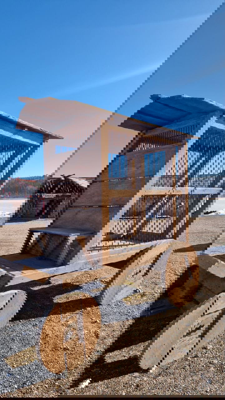 Деревянная тележка с клеткой и массивными круглымі колёсами на гравийной площадке парка; вид на береговую линию и реконструкцию средневекового имущества.