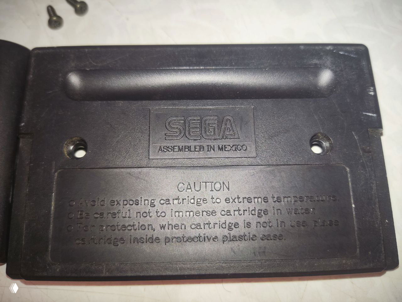 Обратная сторона картриджа с тиснением SEGA и надписью «Assembled in Mexico», видны отверстия под винты, предупреждающая панель и царапины пластика.