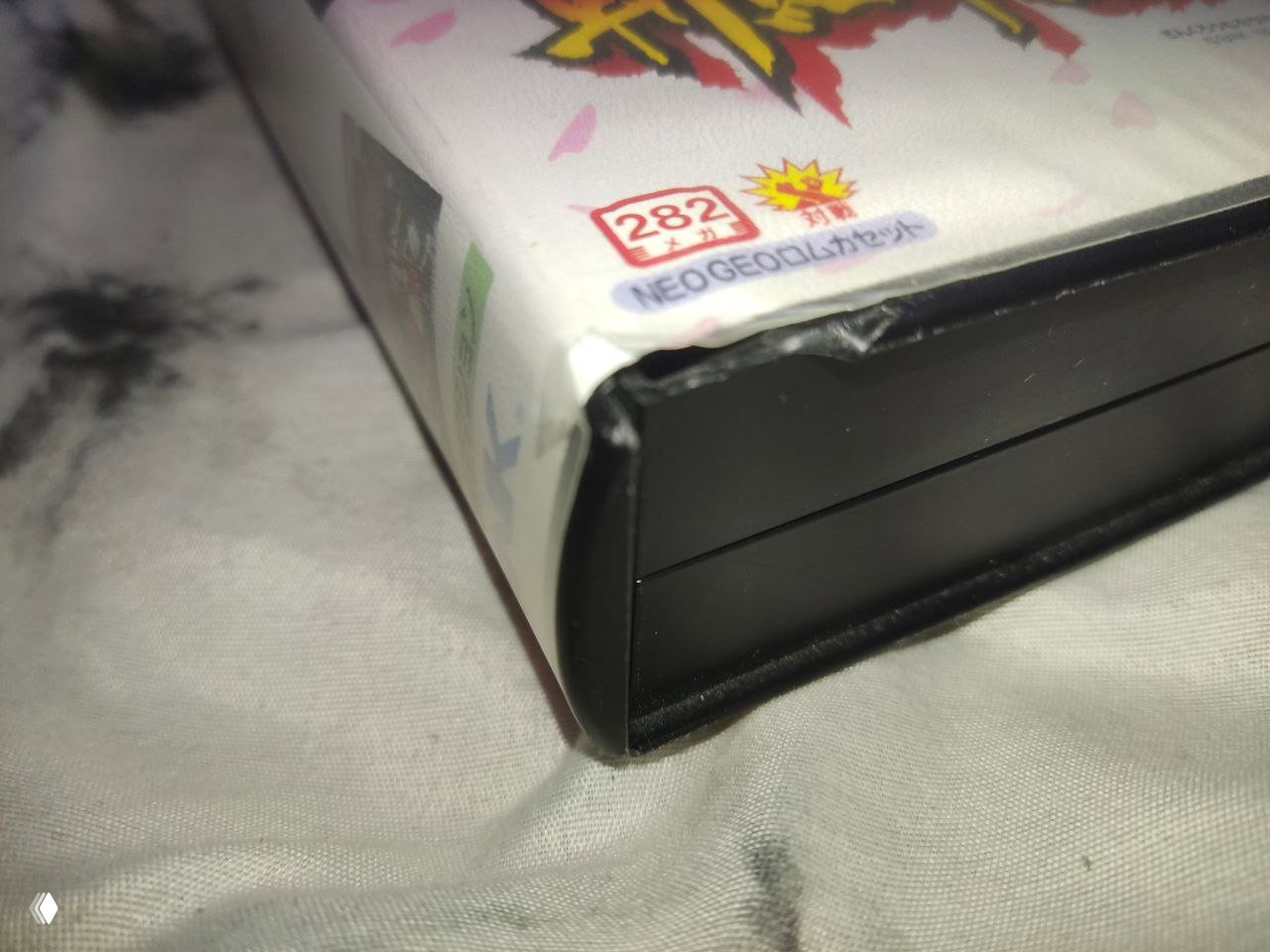 Крупный план повреждённого угла картонной коробки Samurai Spirits 3 для Neo Geo AES — виден продавленный/откушенный угол и защитный пластиковый кейс.