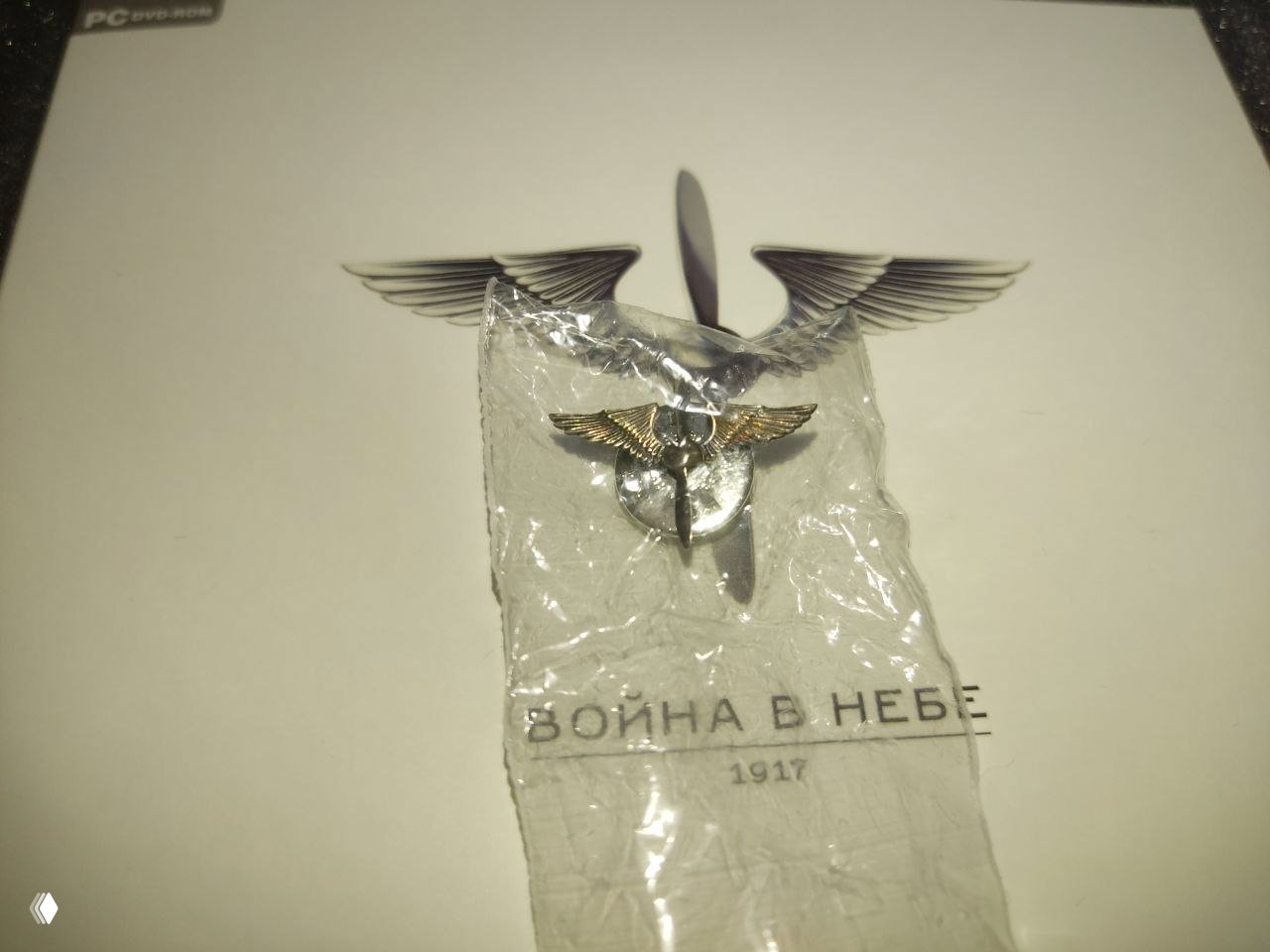 Крупный план металлического значка из набора, упакованного в прозрачный пакетик, на фоне фирменного оформления мануала и коробки.