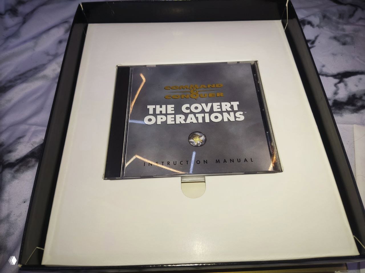 Внутреннее углубление коробки с джевел‑кейсом CD "The Covert Operations" в картонном ложементе, белый держатель диска заметен внизу.
