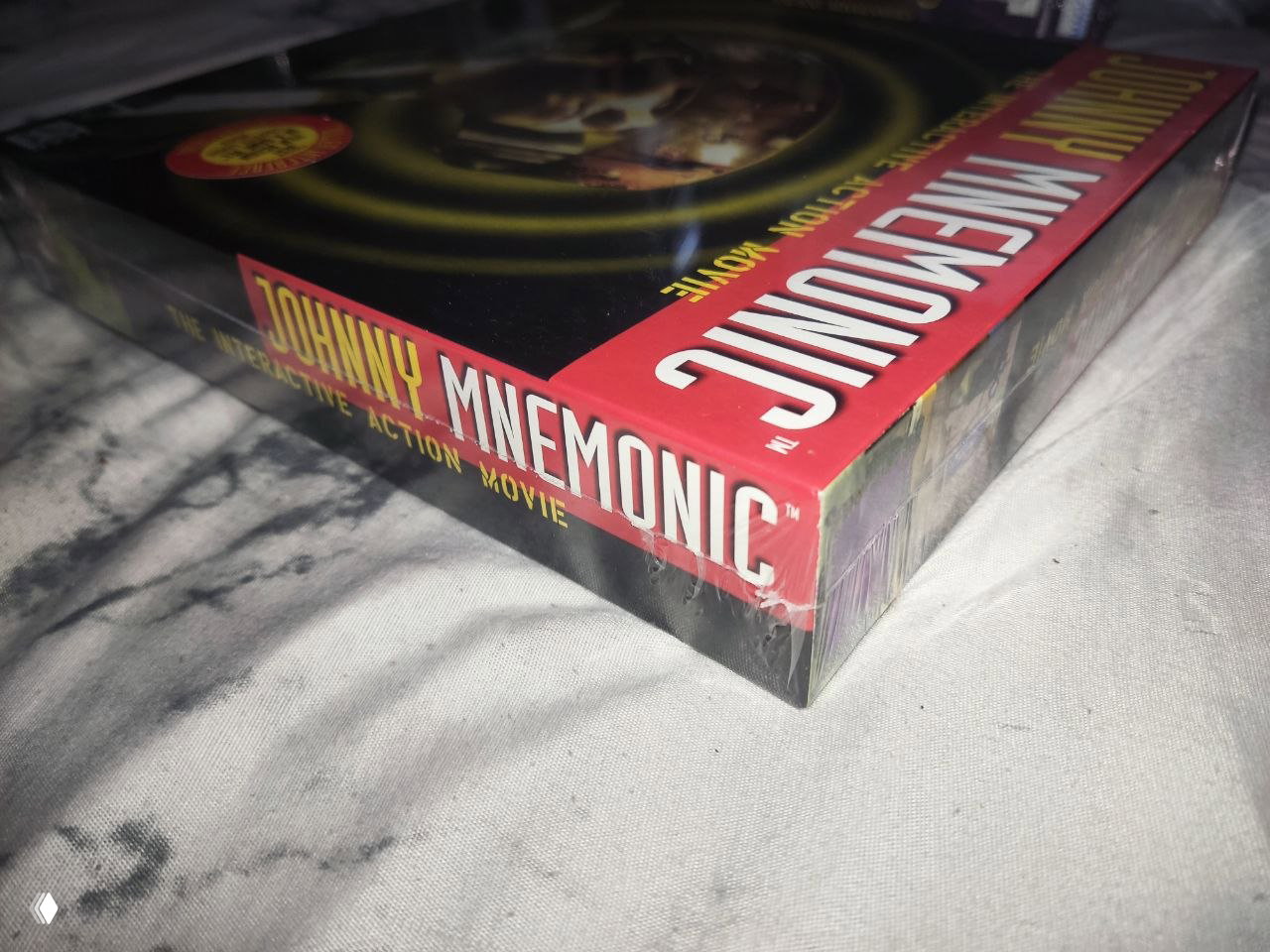 Боковая грань коробки Johnny Mnemonic в плёнке: видно название на торце и часть упаковочной плёнки с небольшими потертостями у краёв.