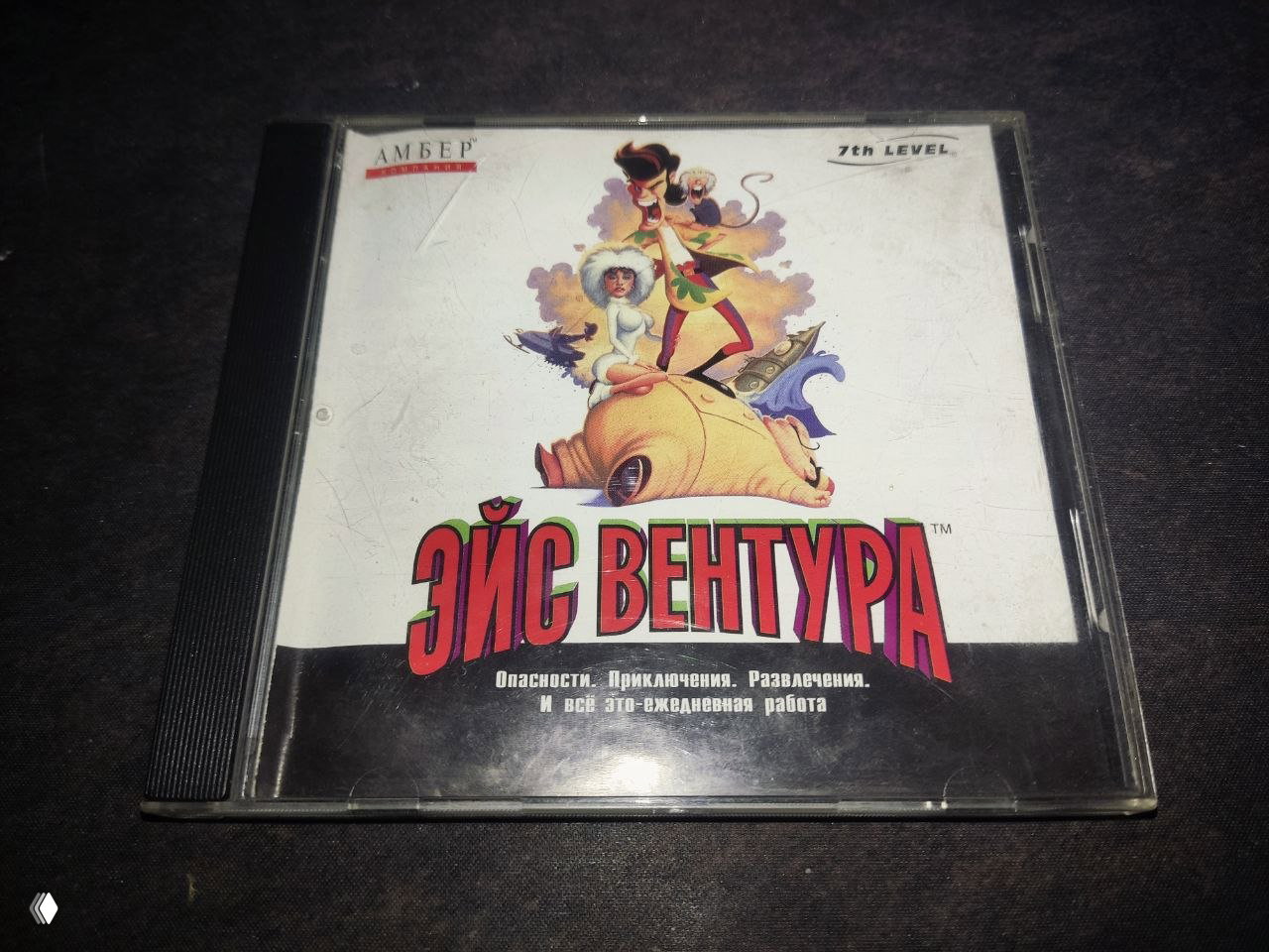 Крупный план коробки игры «Эйс Вентура» в прозрачном боксе на тёмном фоне, видна русская обложка, следы царапин на пластике и чёрный поддон для диска.