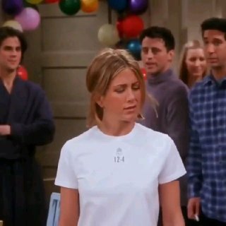 Кадр сцены: женщина в белой футболке смотрит в сторону, на заднем плане несколько людей и разноцветные шарики в помещении.