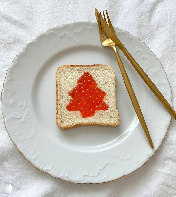 Белая фарфоровая тарелка с кусочком хлеба, на котором выложена красная икра в форме ёлочки; рядом золотые приборы, вариант лёгкого ужина.