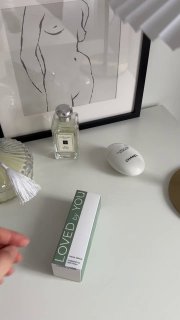 Фотография упаковки Loved by You на белом столе рядом с флаконом духов и постером с линейным силуэтом в уютном интерьере.