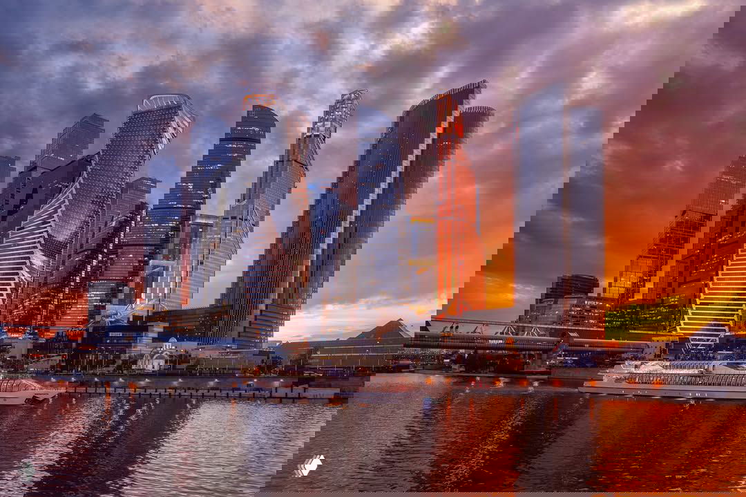 Силуэты небоскрёбов Москва‑Сити на закате над рекой, отражение в воде и прогулочный теплоход у берега.