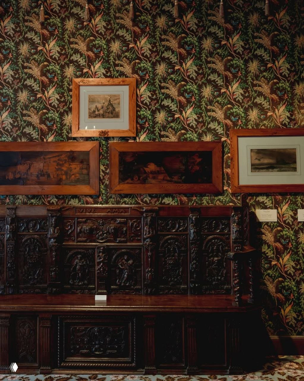 Резная деревянная скамья и картины на фоне растительного обоев, тёмная мебель и декоративная панель — одна из комнат дома Виктора Гюго с богатой отделкой.