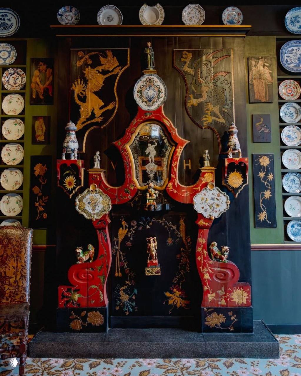 Декоративный красный камин с фарфоровыми элементами и фигурками, окружённый стеной из тарелок и барельефов — центральный экспонат интерьерного зала.