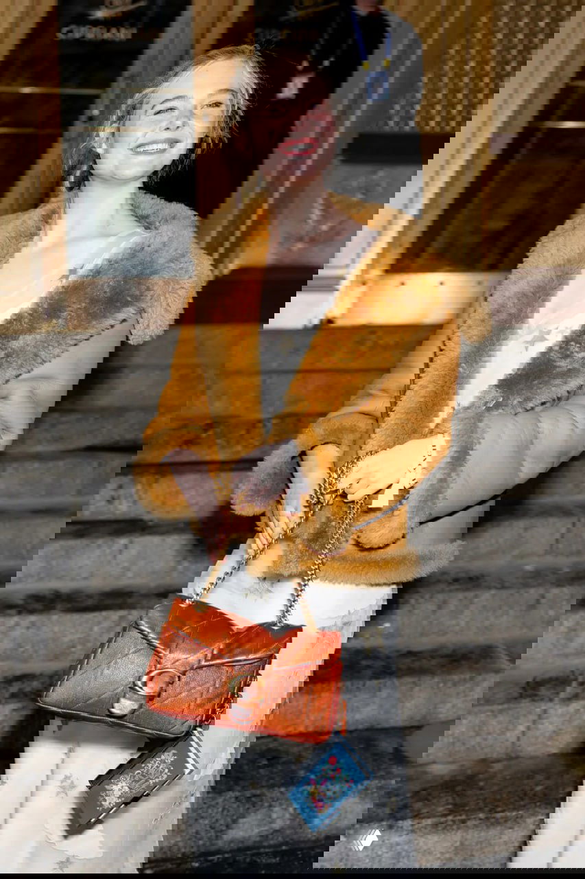 Женщина с сумкой носит миниатюрную книгу-брелок Coach: видна подвеска-книга, прикреплённая к цепочке сумки на уличном кадре.