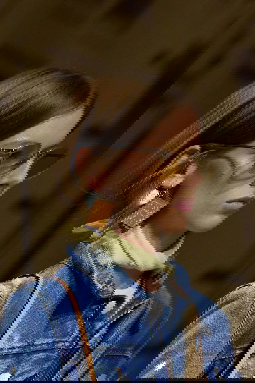 Крупный портрет модели в коричневой куртке с серьгой-миникнигой и солнечными очками, акцент на украшении в виде книги.