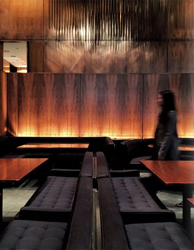 Тёмный уютный салон ресторана с низкими диванами и деревянными панелями на стенах; силуэт человека в движении подчёркивает световое решение интерьера и интимность пространства.